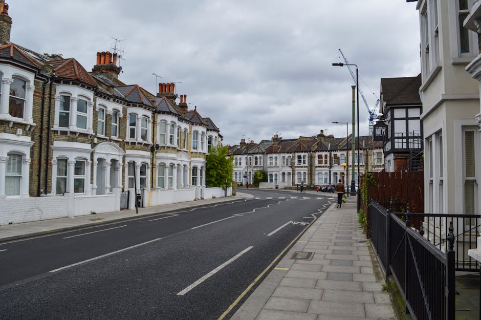 A residential street scene featuring a row of Victorian-style terraced houses with white facades, bay windows, and red-tiled roofs, situated along a paved sidewalk with iron railings. The street is em
